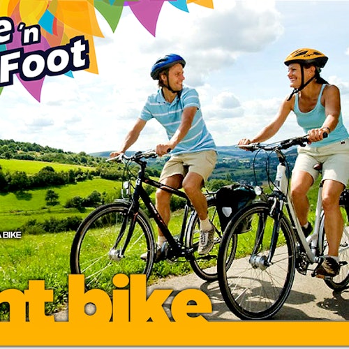 A man and a woman riding bikes on a scenic path, wearing helmets and casual attire, with green hills and cloudy sky in the background.