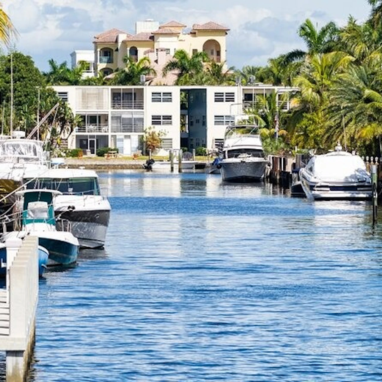 A canal lined with boats and modern houses surrounded by palm trees under a sunny, partly cloudy sky.