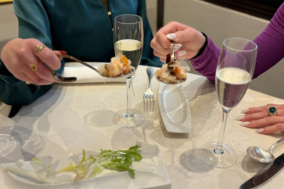 Two people dining, holding spoons with food over a table set with plates, greenery, and glasses of sparkling wine.