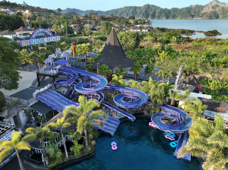 A water park featuring purple water slides, a pool, tropical plants, and a scenic background of hills and a lake.
