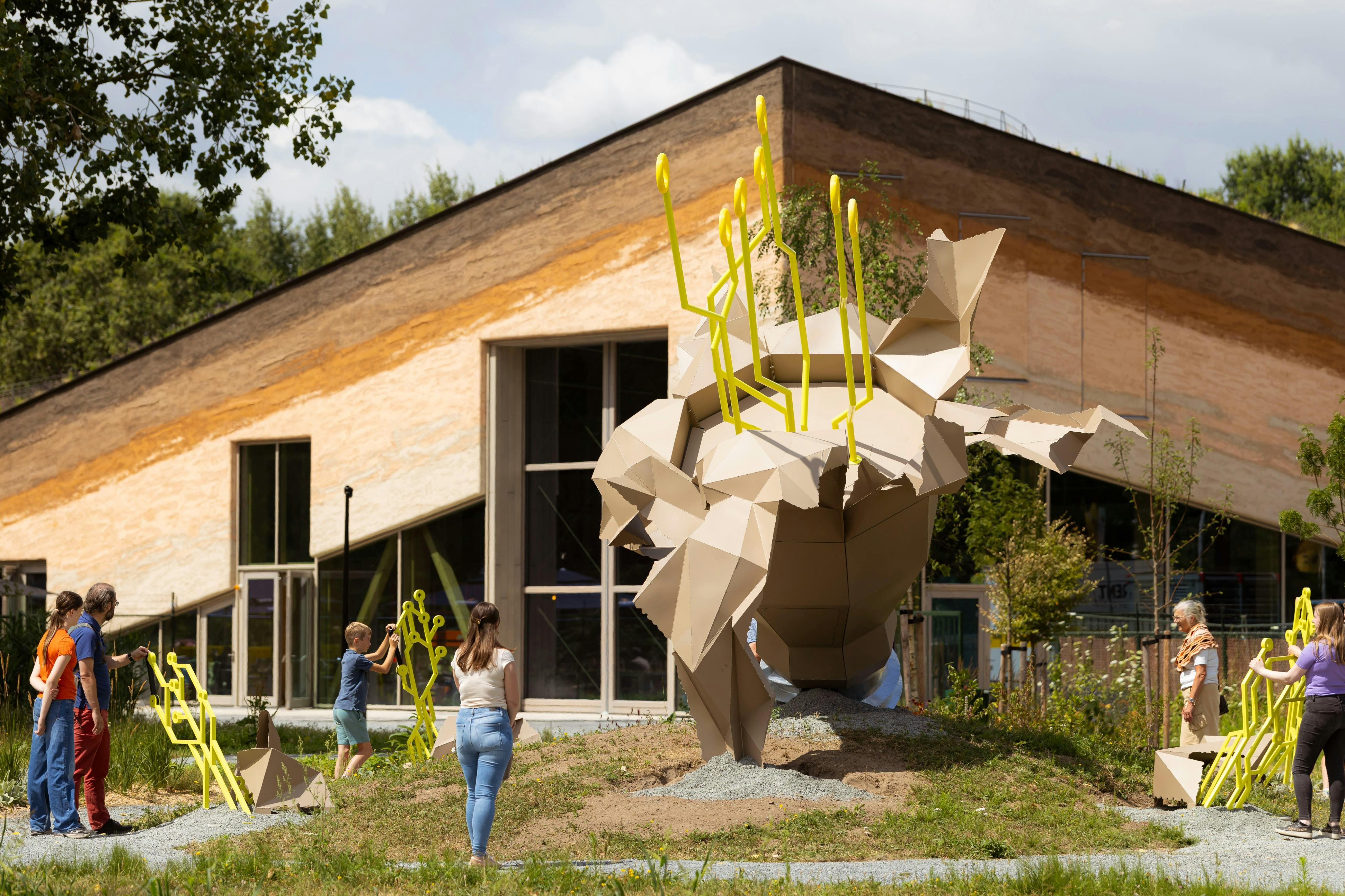 Una escultura geomètrica amb varetes grogues en un entorn de jardí, envoltada de gent prop d'un edifici modern amb sostre inclinat.