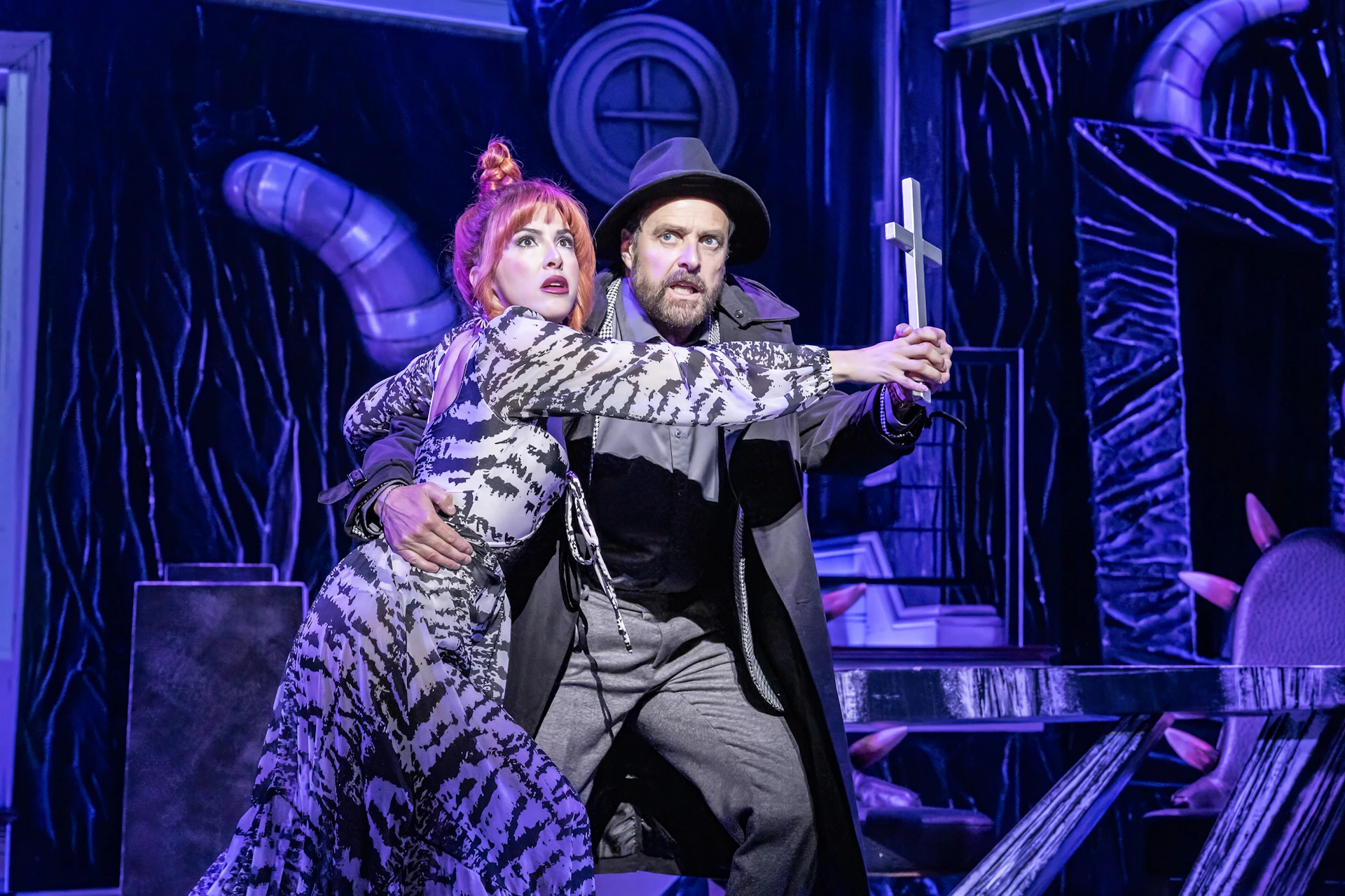Two people on stage in dramatic poses, one holding a cross. The setting features theatrical lighting and dramatic props.