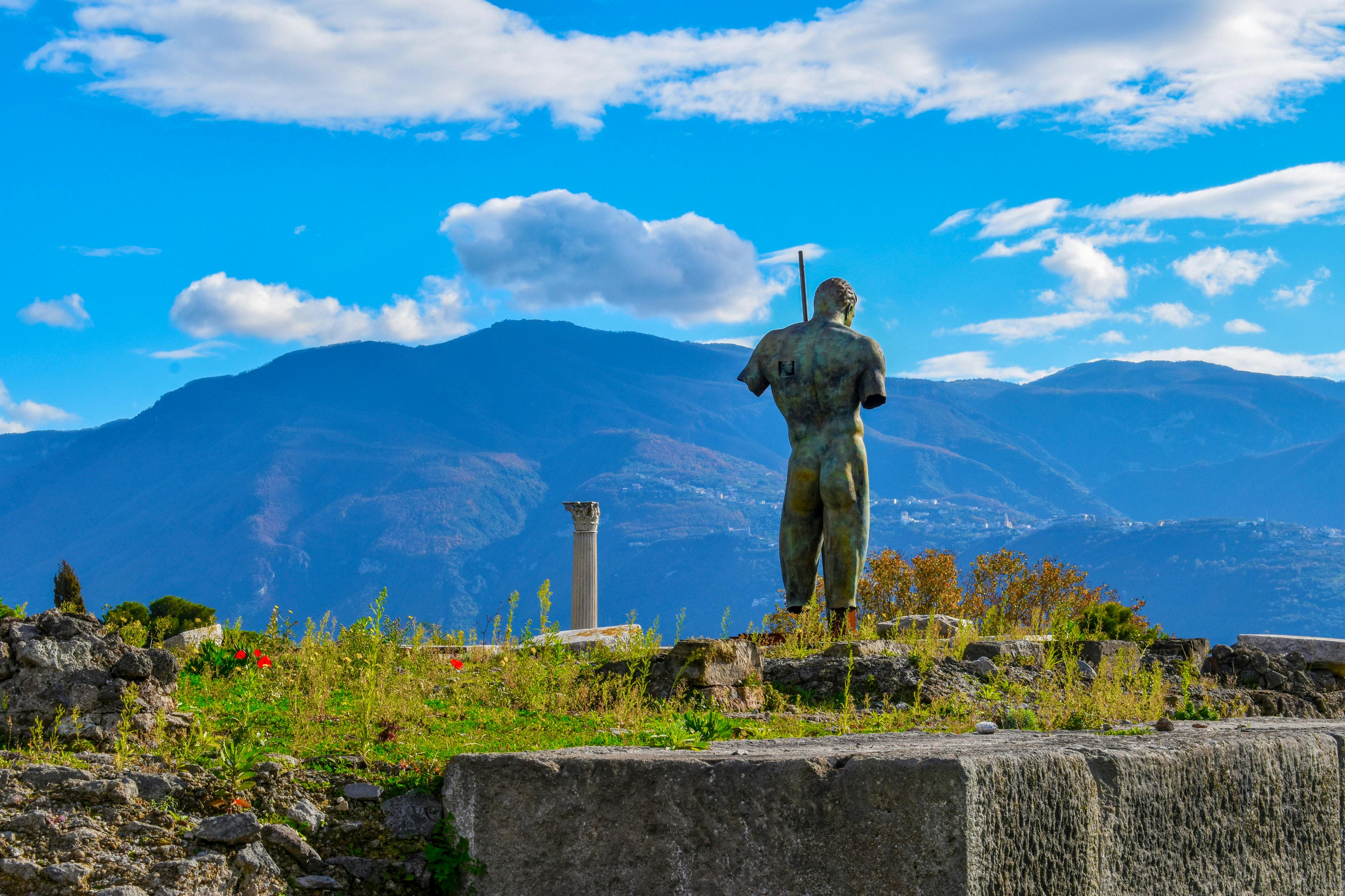 古代遺跡の上には向かい合った像が立っており、雲が散在する明るい青い空の下には、山々と1本の柱が見えます。