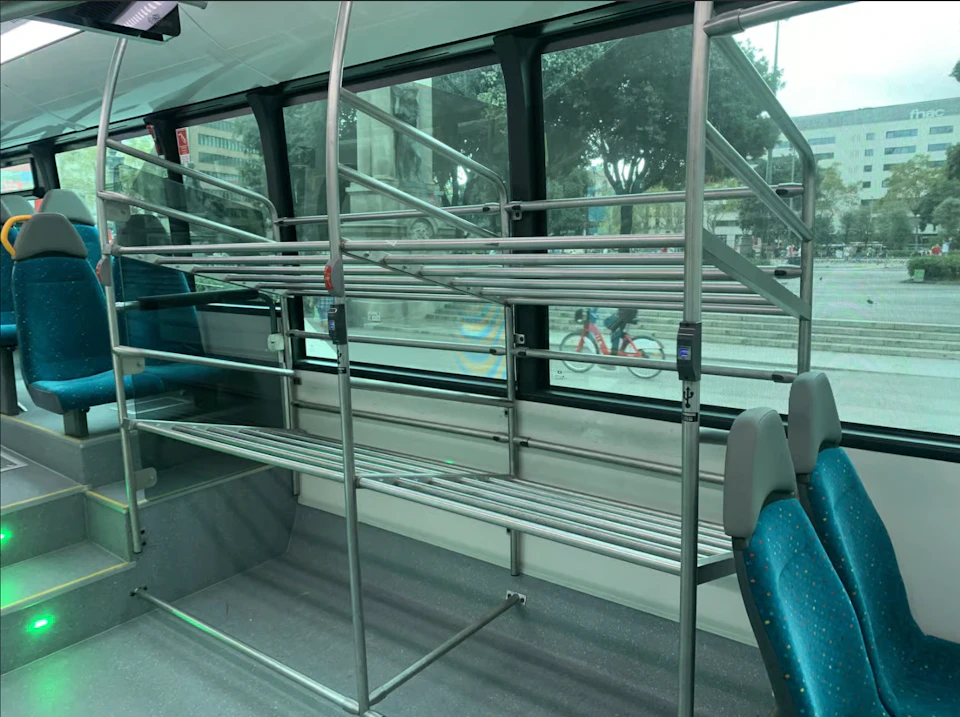 Interior of a bus with metal luggage racks, blue-patterned seats, and a view of city streets through large windows.