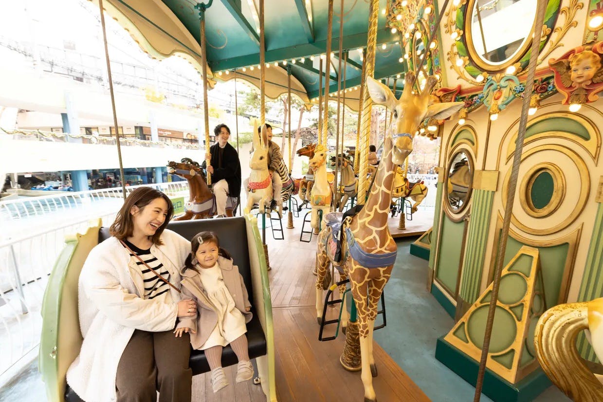 一名妇女和儿童坐在旋转木马长凳上，周围围绕着长颈鹿和兔子等动物形象，背景中还有其他骑手。