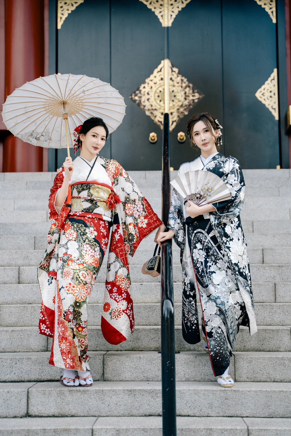Two women dressed in traditional kimonos, holding a parasol and a fan, stand on steps in front of an ornate doorway.