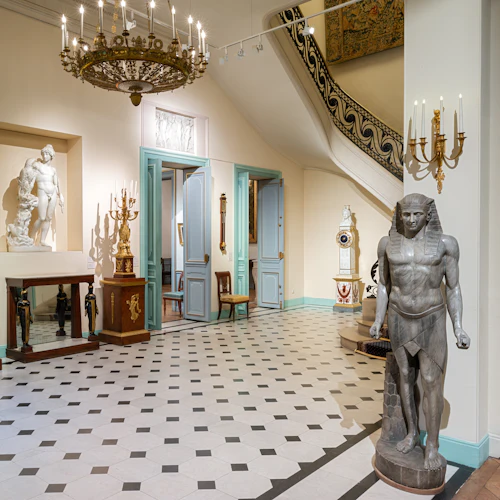 Museuminteriör med egyptiska statyer, klassiska skulpturer, utsmyckade ljuskronor och antika möbler i ett kaklat rum med ljusblå dörrar.