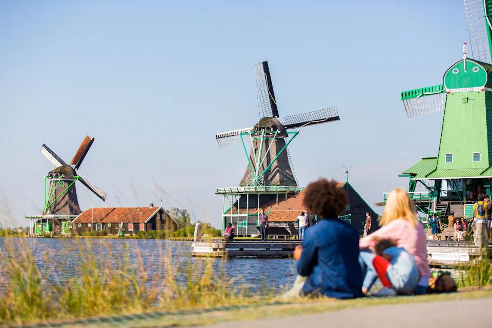 People sit along a canal, with traditional windmills and buildings in the background under a clear blue sky.