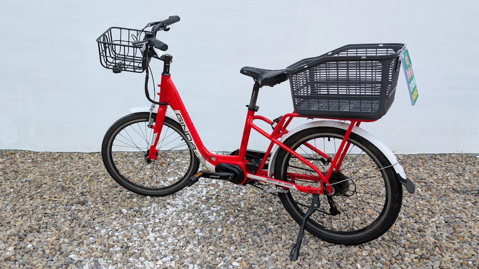 A red bicycle with a front basket and a large rear cargo basket, parked on a gravel surface against a white wall.