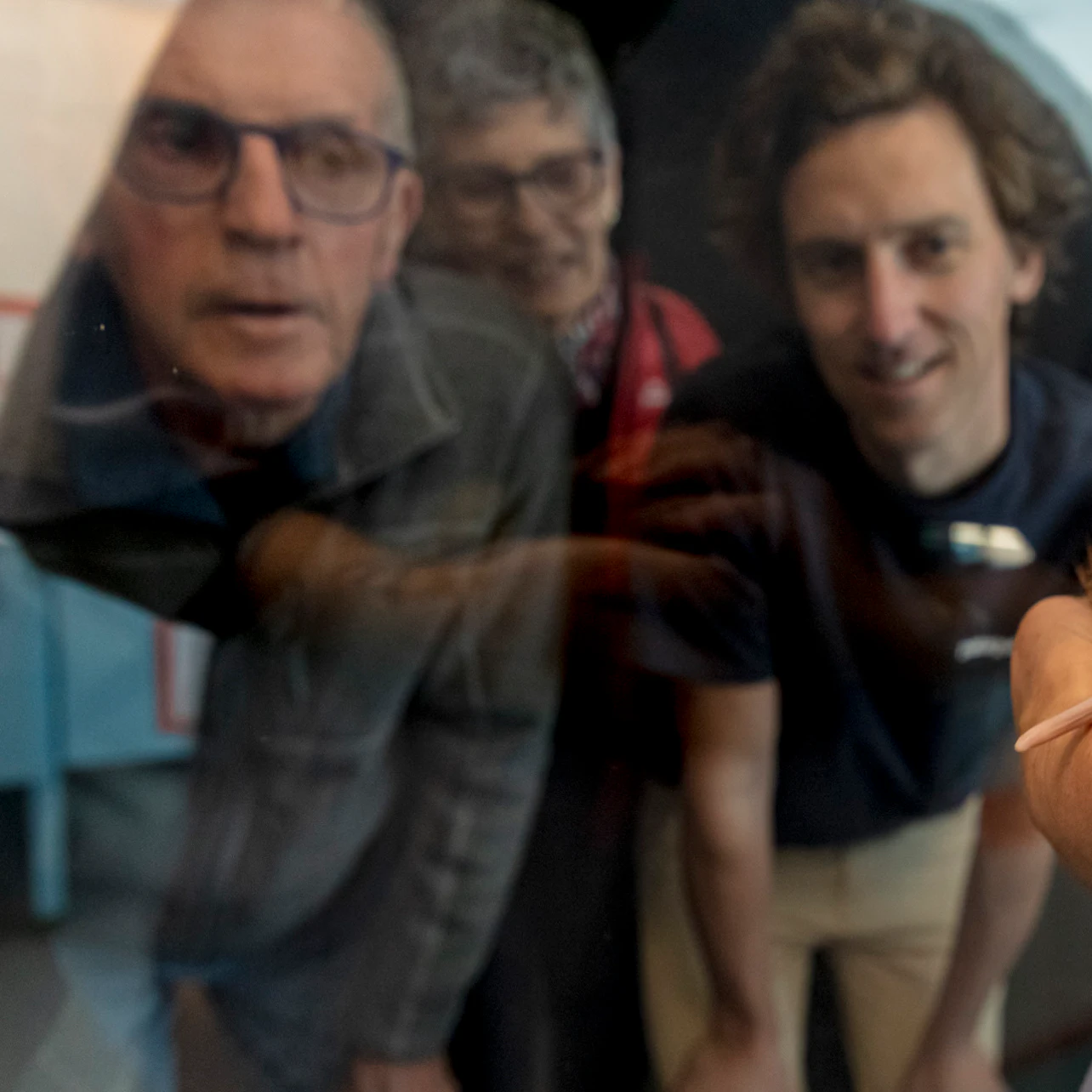 Three people looking through a reflective surface while a hand holds a small object in the foreground of the image.