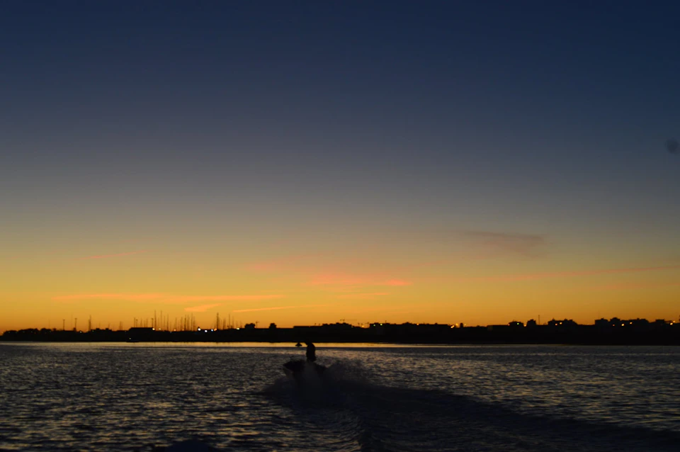 A silhouette of a person on a jet ski rides on a calm sea during sunset, with a distant coastline and boats in the background.