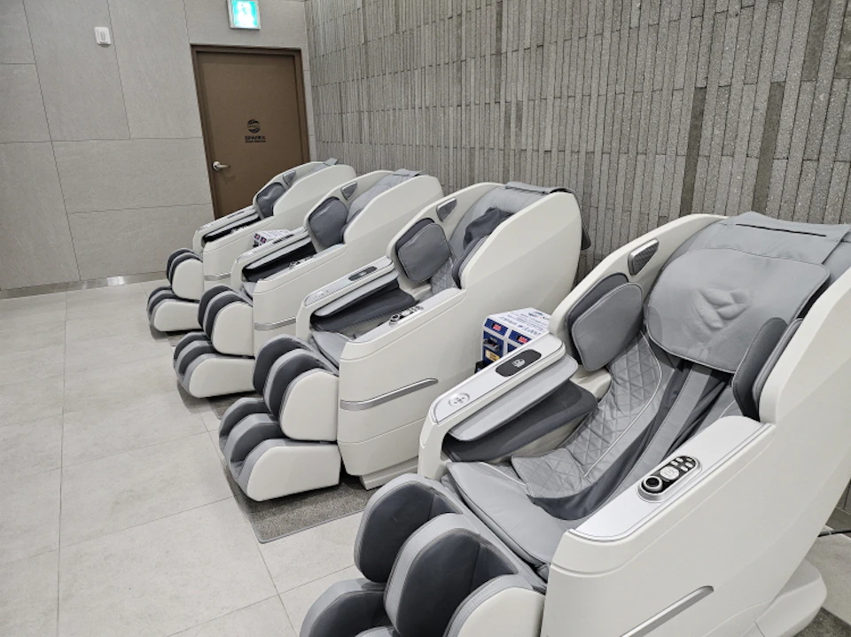 A row of modern white massage chairs with gray padding in a clean indoor room next to a wooden door labeled in the background.