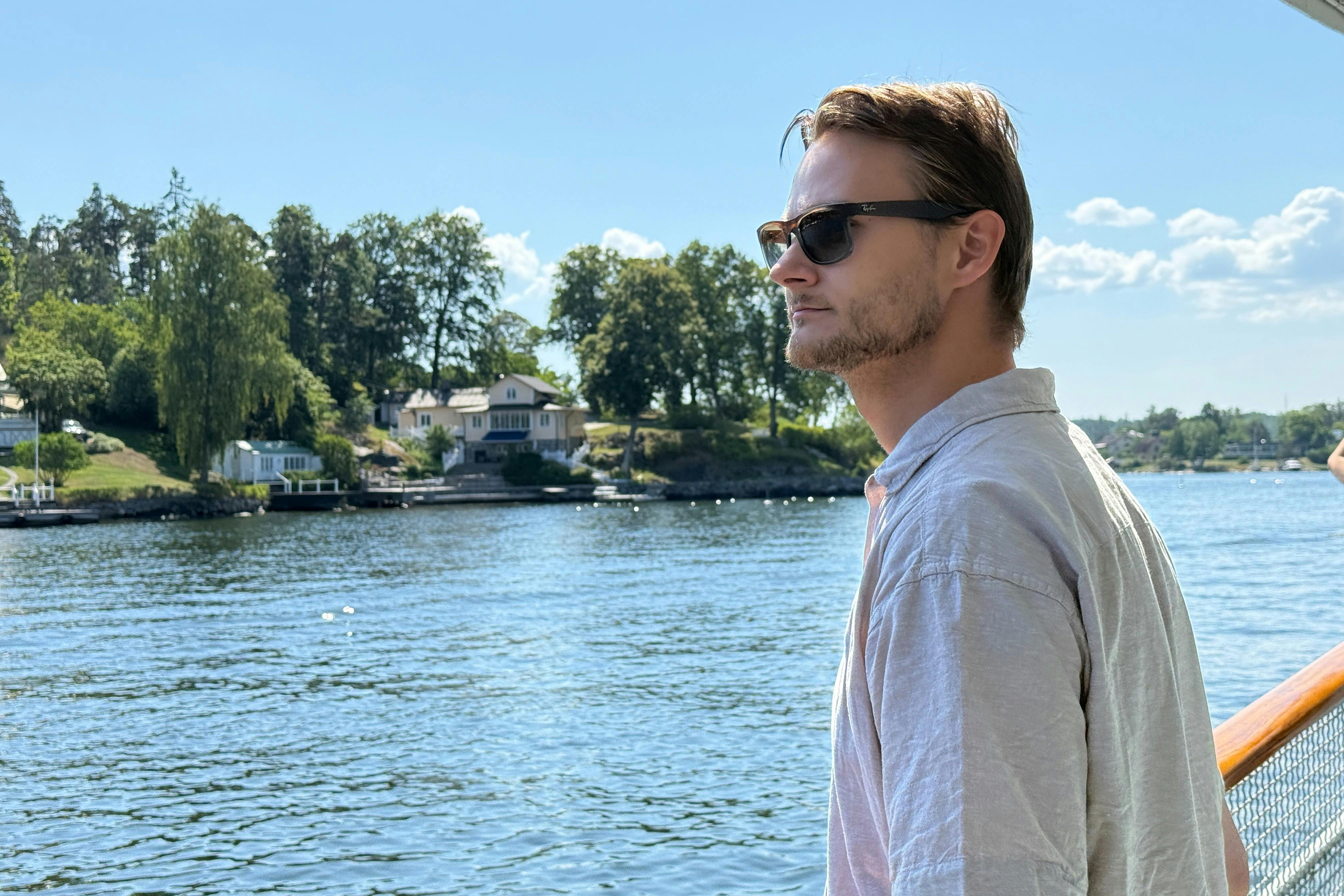 Uomo con occhiali da sole guarda l'acqua da una barca, con case, alberi e un cielo parzialmente nuvoloso sullo sfondo.