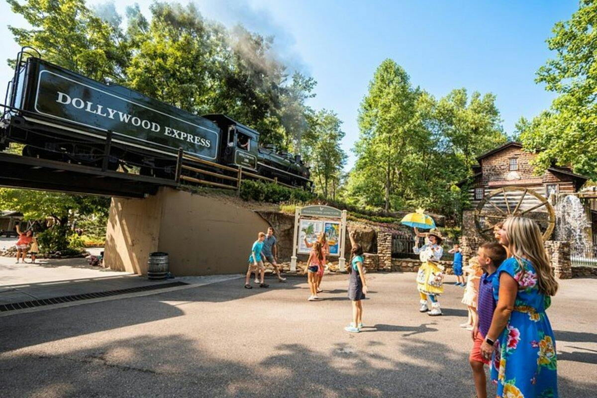 A steam train labeled 'Dollywood Express' passes over a bridge while people enjoy the area below, including a performer.