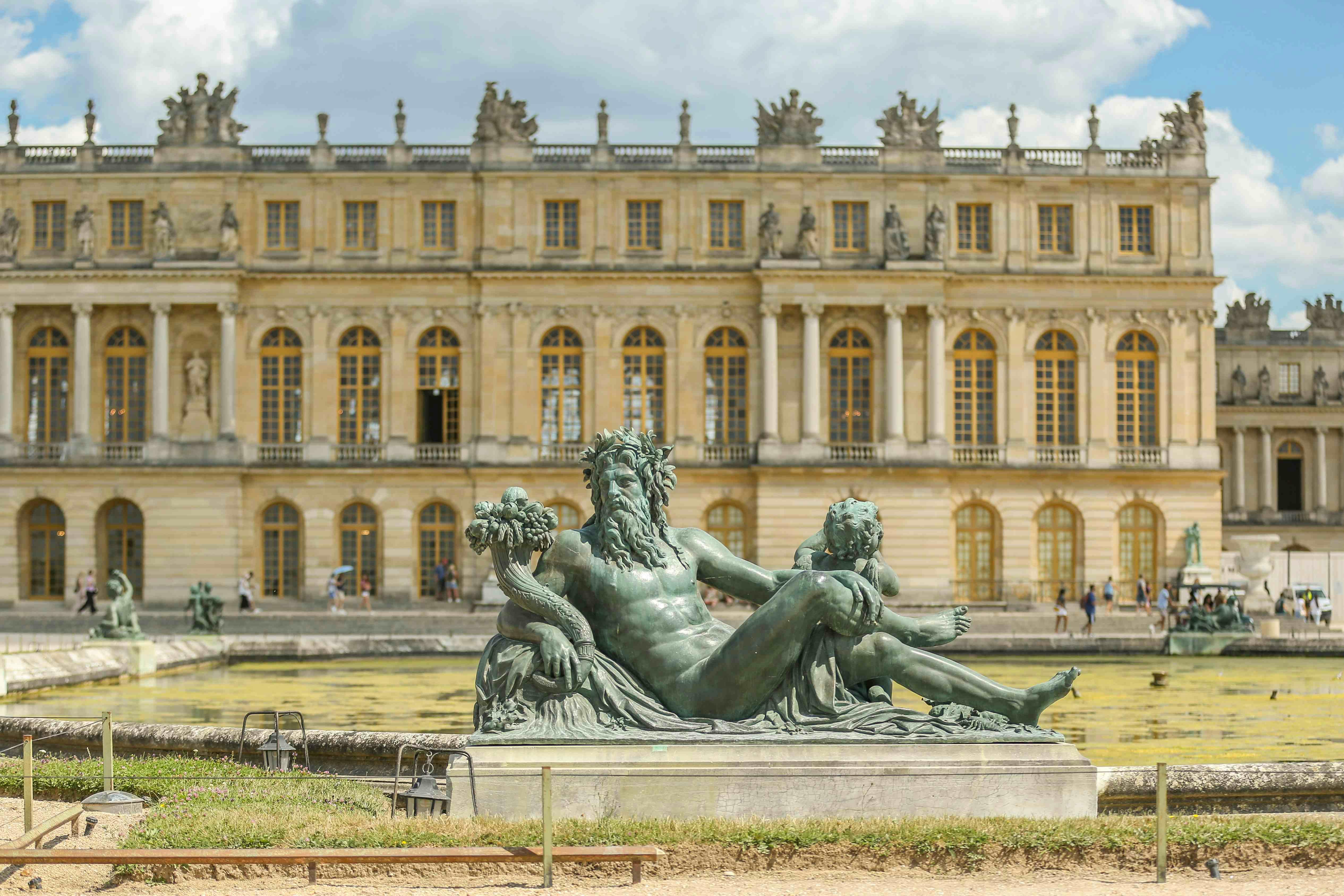 A sculpture of a reclining man with a child near an ornate building with numerous windows, on a sunny day, with a few people in the background.