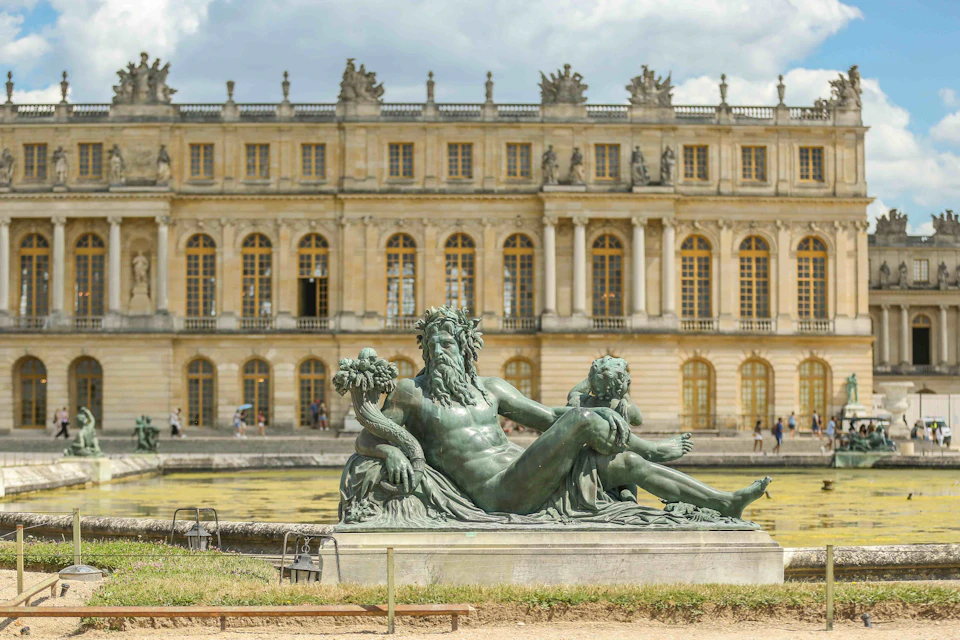 A sculpture of a reclining man with a child near an ornate building with numerous windows, on a sunny day, with a few people in the background.