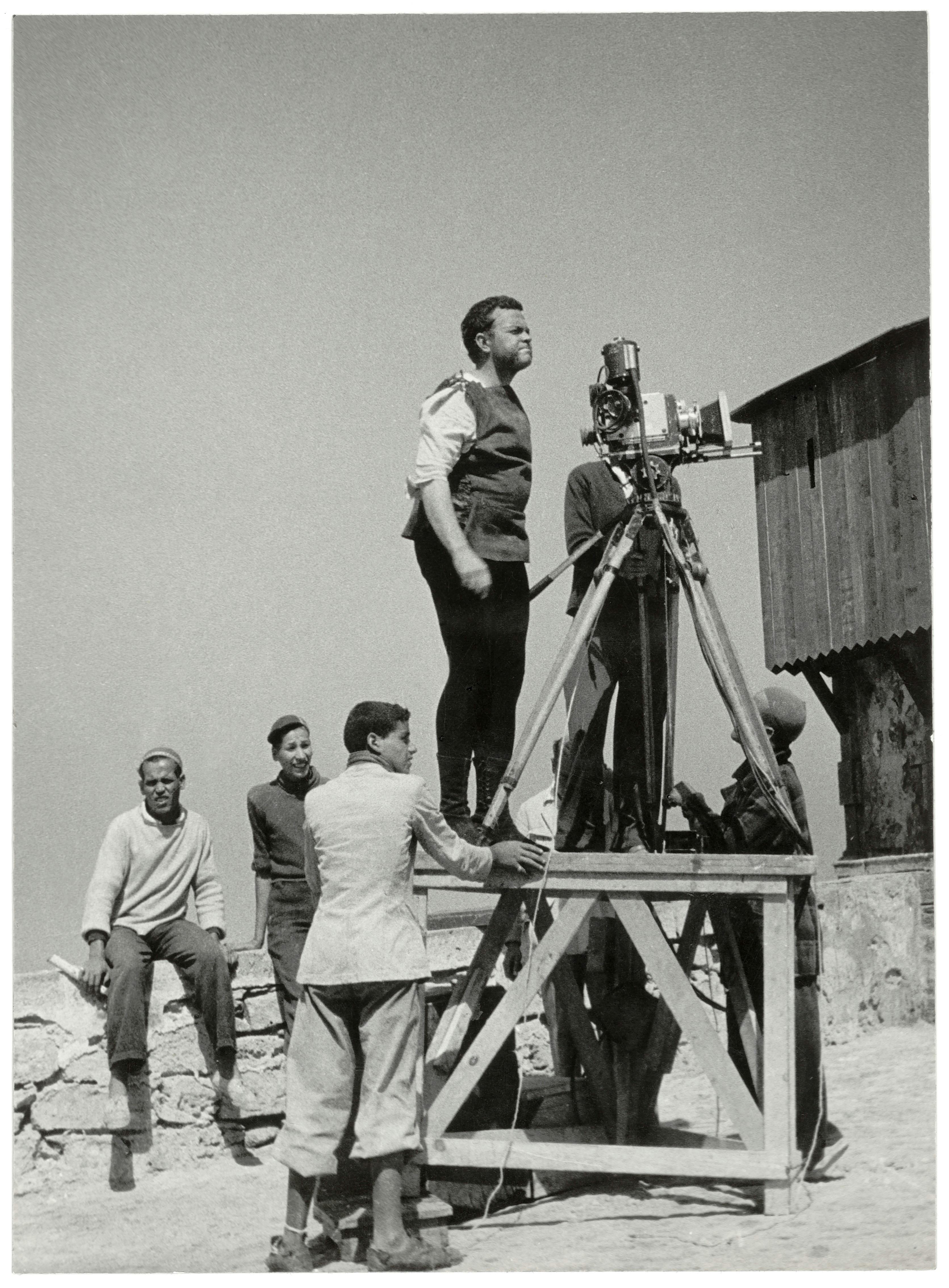 Un hombre está de pie sobre una plataforma de madera manejando una cámara cinematográfica, con varios otros observando o ayudando cerca.