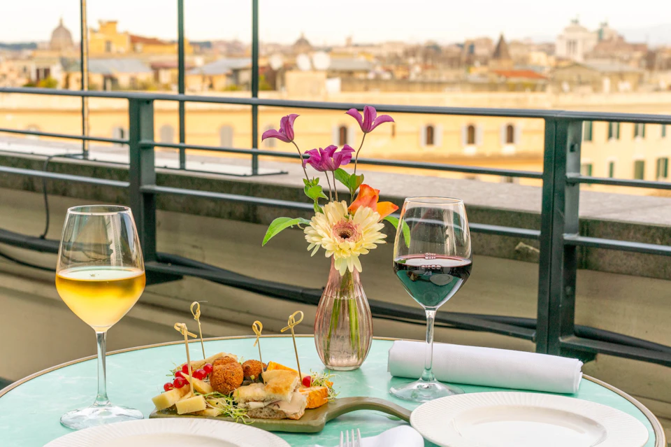 A rooftop table with wine glasses, a vase of flowers, and appetizers, overlooking a cityscape in the distance.