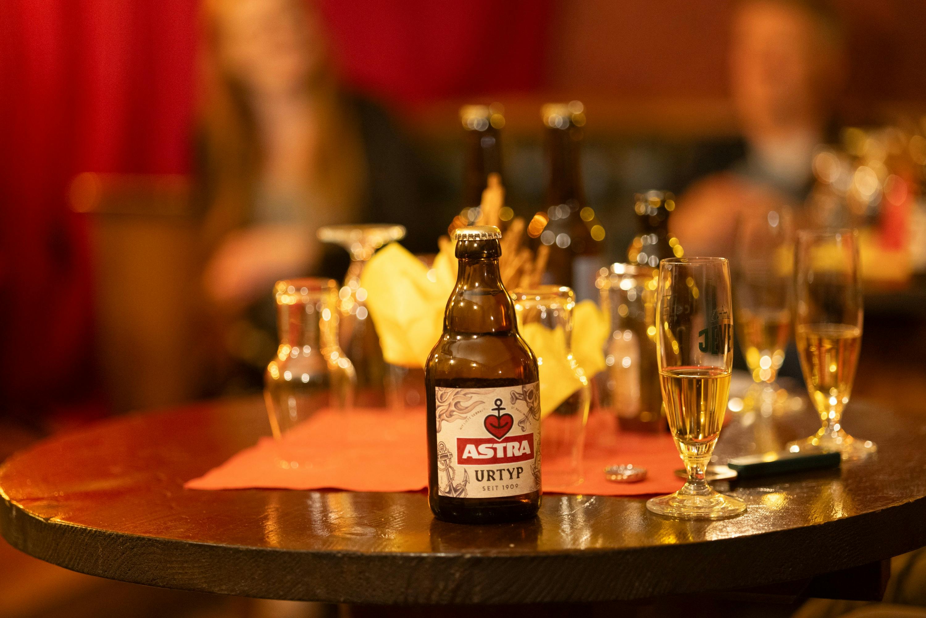 Flaske Astra øl på et bord med forskellige glas og flasker i en svagt oplyst indstilling; sløret figur i baggrunden.