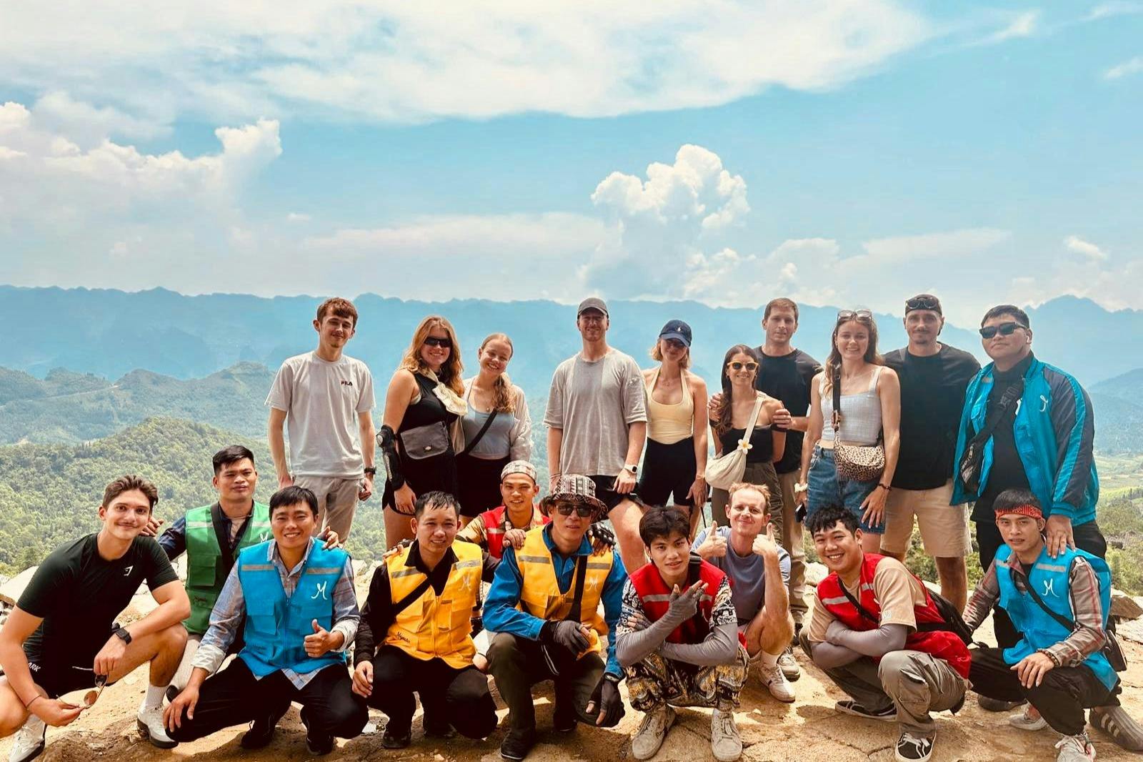 En gruppe på tretten personer, der poserer ved et bjergudsigtspunkt med frodige grønne bakker og en delvist overskyet himmel i baggrunden.
