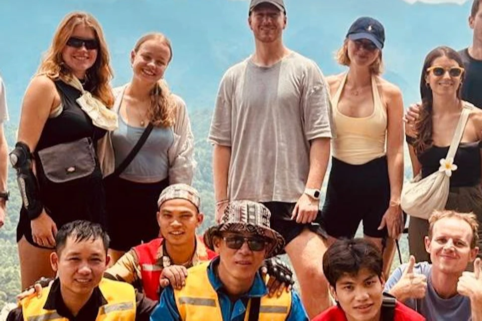 A group of thirteen people posing at a mountain viewpoint with lush green hills and a partly cloudy sky in the background.