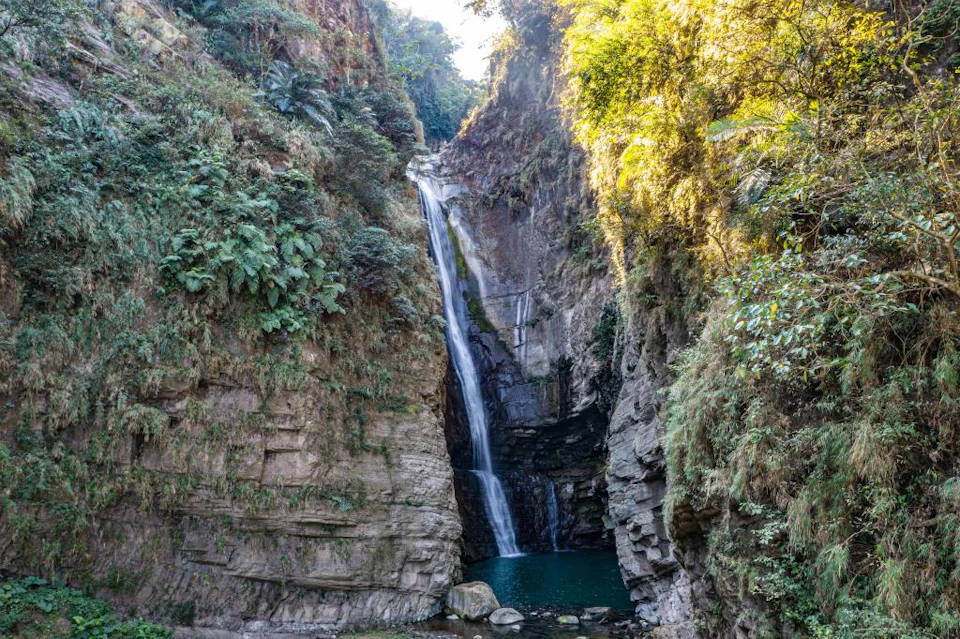 A waterfall cascading down a rocky cliff surrounded by lush green vegetation into a small pool below.
