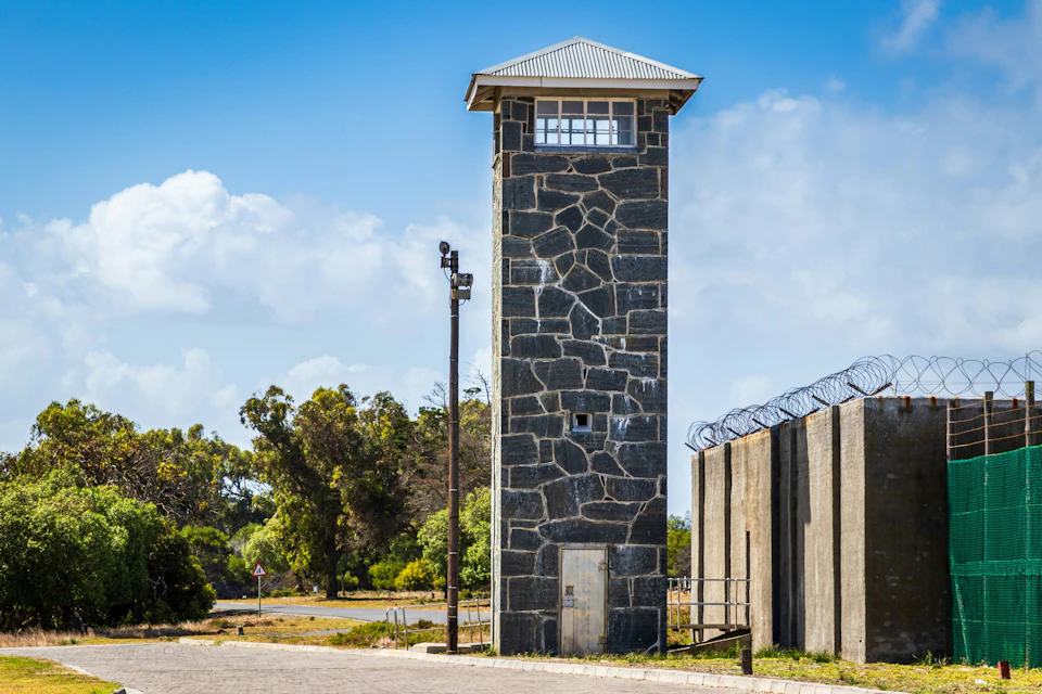 Tall stone watchtower with a peaked roof next to a barbed-wire fence, with trees and a clear blue sky in the background.