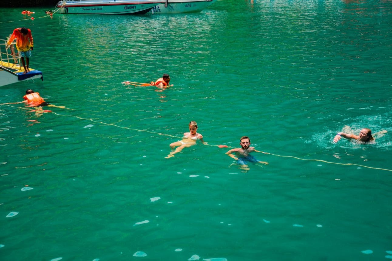 Αρκετοί άνθρωποι κολυμπούν σε καθαρά πράσινα νερά κοντά σε μια βάρκα, με κάποιους να κρατιούνται από ένα πλωτό σχοινί.