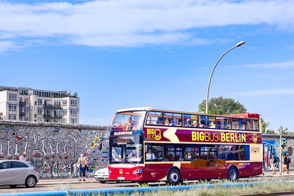 A red double-decker sightseeing bus labeled 'Big Bus Berlin' drives past a graffiti-covered wall on a sunny day.