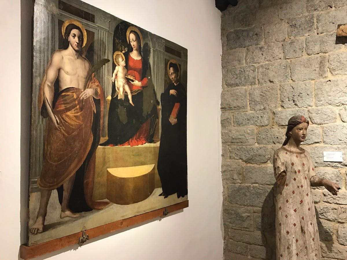 Pokój z obrazem religijnym na ścianie i kamiennym posągiem kobiety przy ścianie z cegieł.