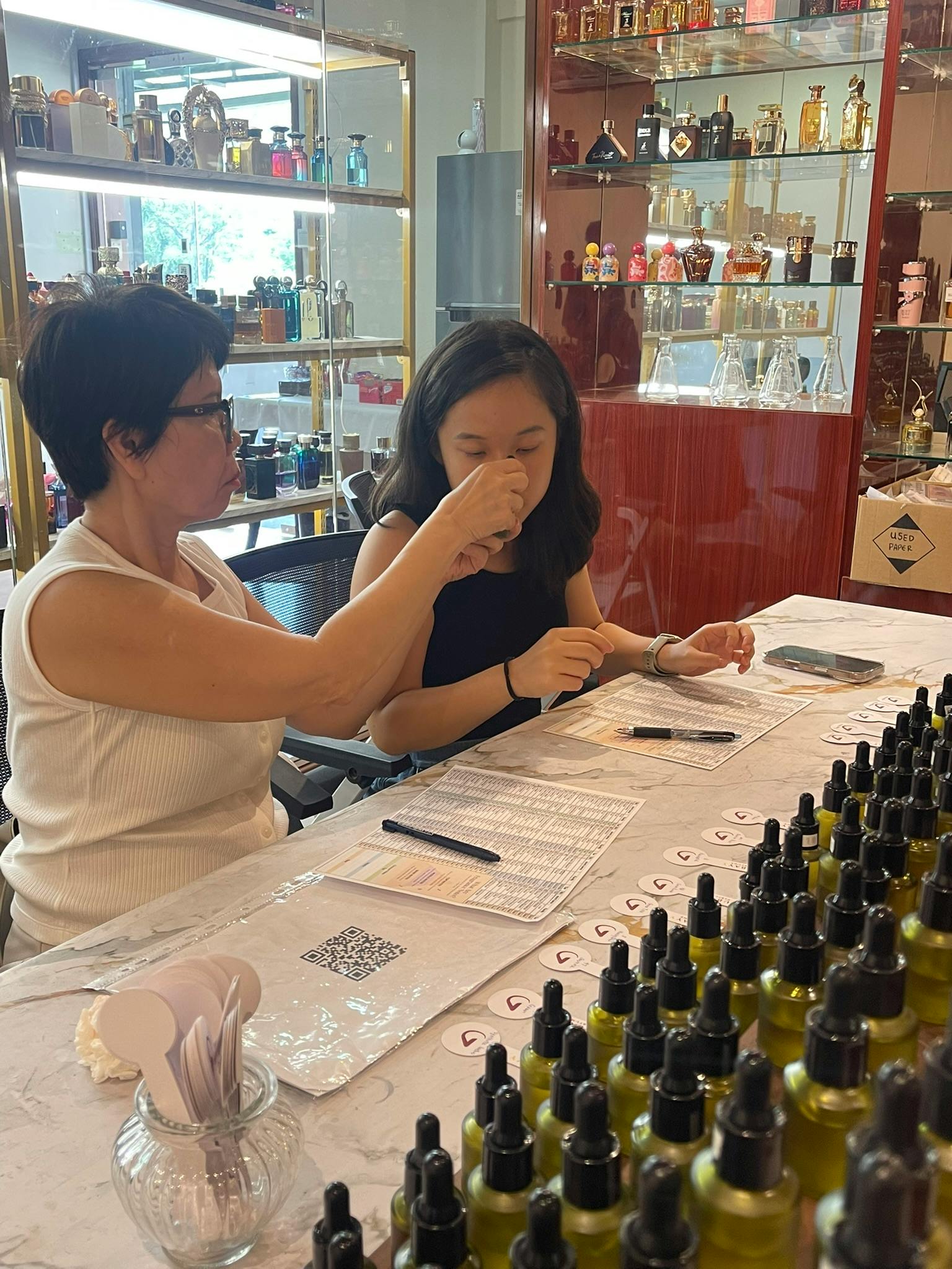 Two people seated at a table in a perfume shop. One is smelling a fragrance strip, with rows of bottles in front of them.