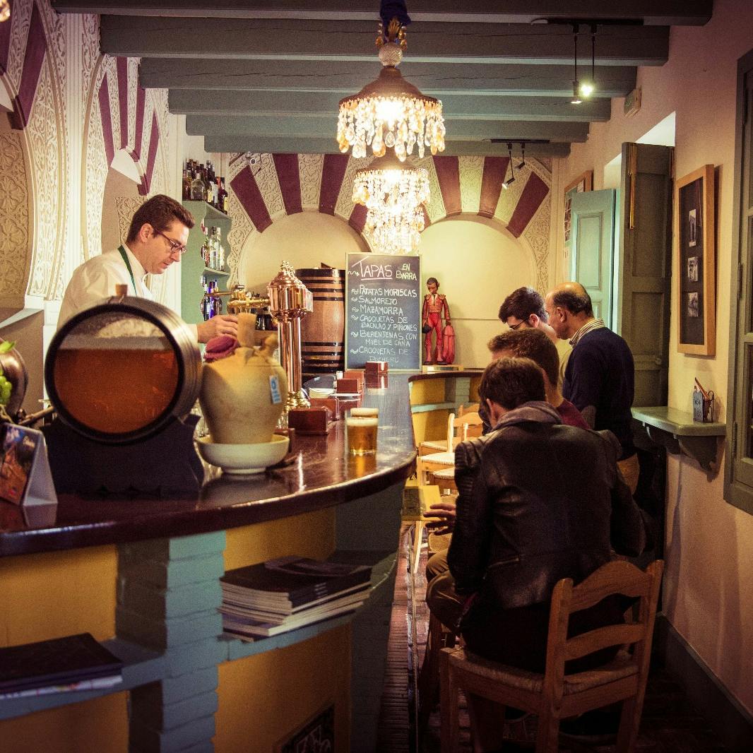 人们坐在舒适的小吃酒吧里，调酒师提供饮料、华丽的吊灯和黑板菜单。