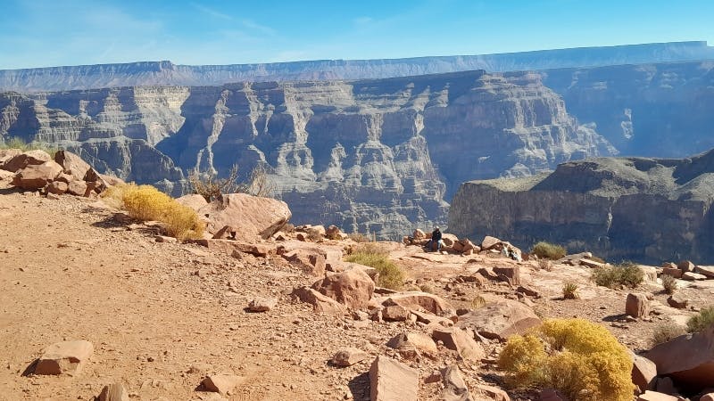 A vast canyon landscape under a clear sky, with rocky terrain and two people sitting among boulders in the foreground.