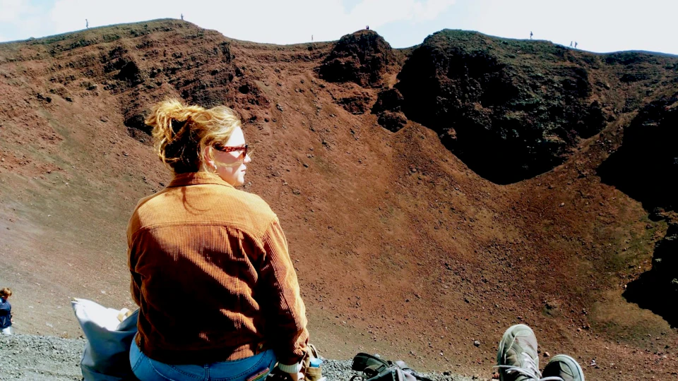 Person with sunglasses in brown jacket stands in front of a rocky, red-brown hillside under a partly cloudy sky.