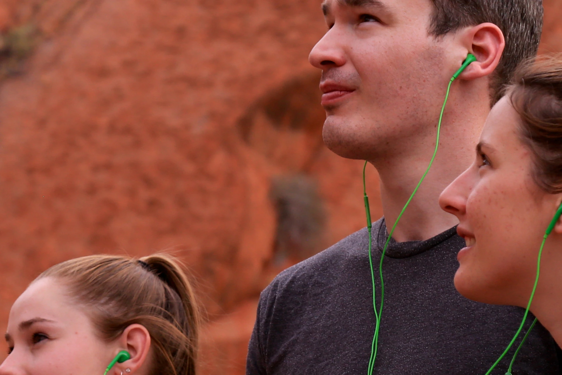 Three people wearing earphones, holding smartphones, and looking up, standing in front of a large, textured rock formation.
