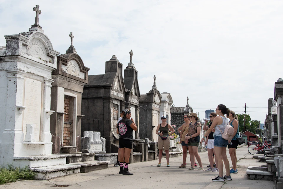 A group of people on a guided tour in a cemetery with above-ground tombs on a cloudy day.