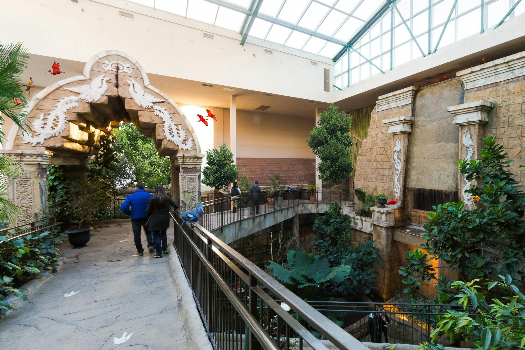 Indoor garden with a stone archway, plants, and decorative columns. People walk on paths surrounded by greenery. Red birds fly above.