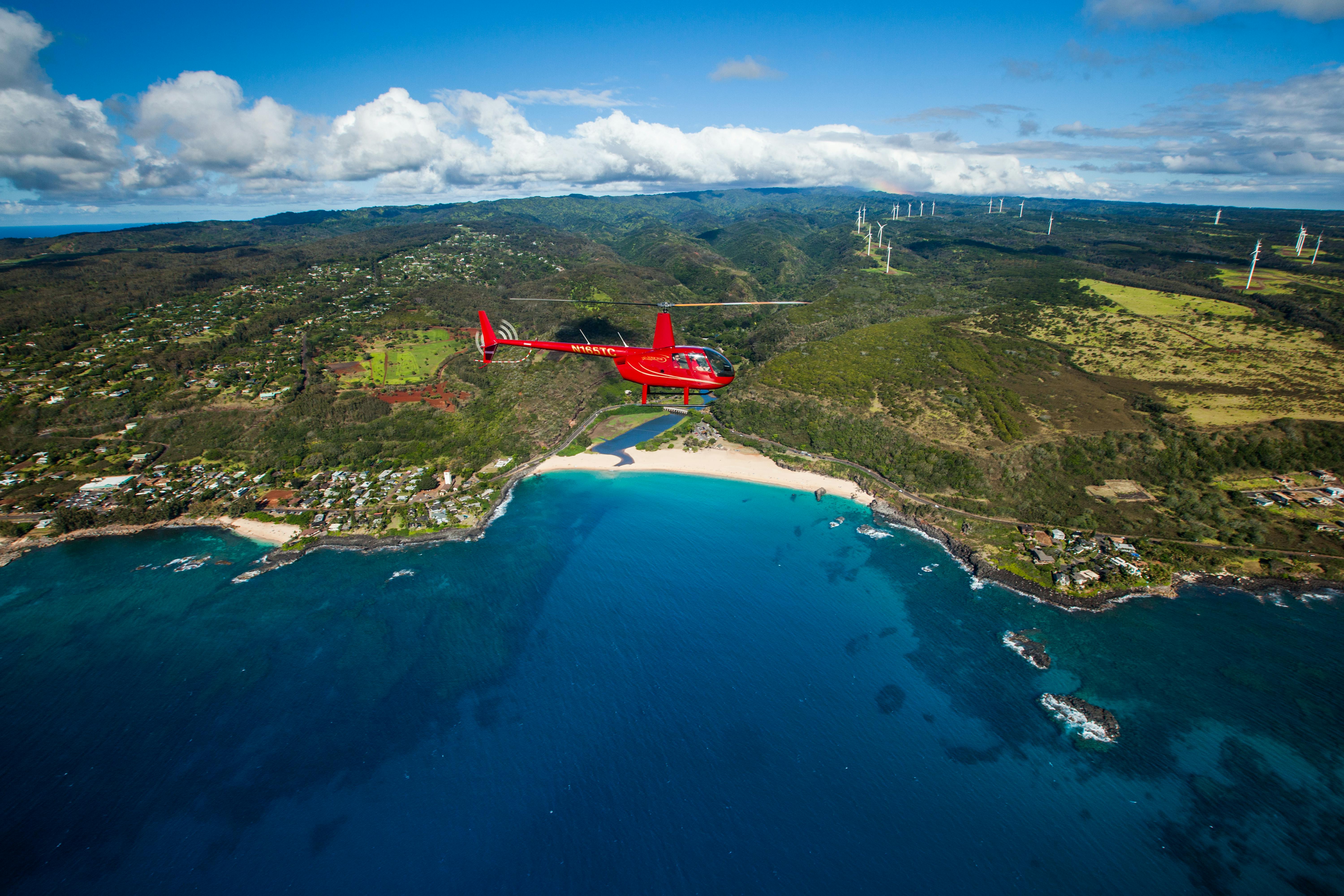 A red helicopter flies over a coastal landscape with turquoise waters, a sandy beach, houses, hills, and distant wind turbines.