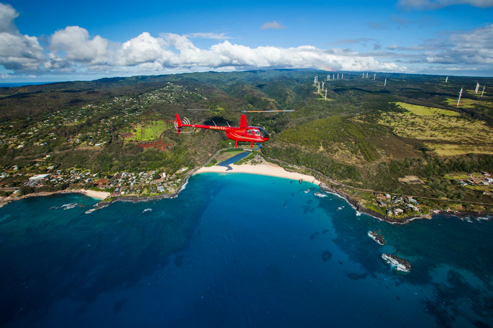 A red helicopter flies over a coastal landscape with turquoise waters, a sandy beach, houses, hills, and distant wind turbines.