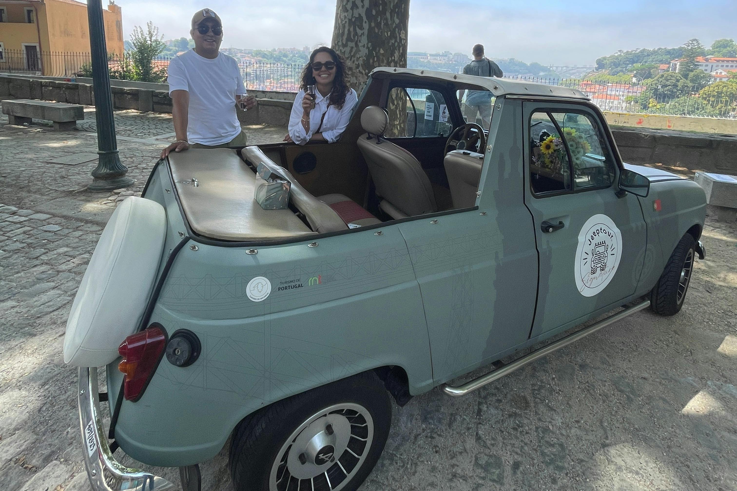 Two people stand and sit by an open-top vintage car parked on a cobblestone street near a city overlook.