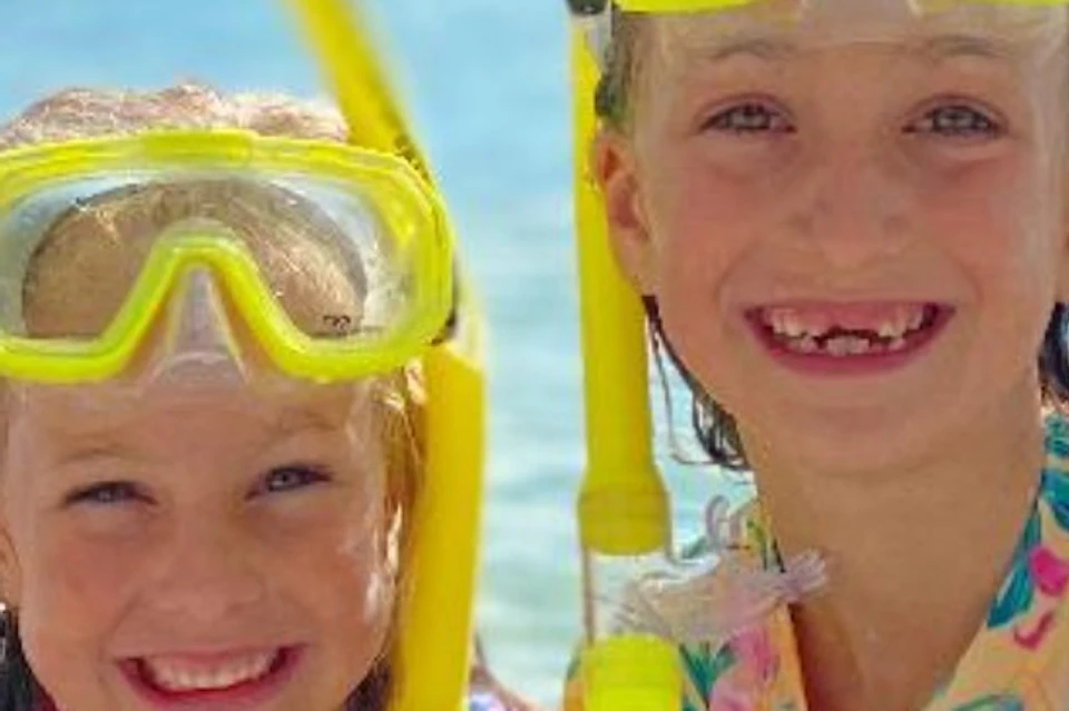 Two children wearing colorful swimsuits and yellow snorkeling gear, smiling in shallow, clear water under a sunny sky.