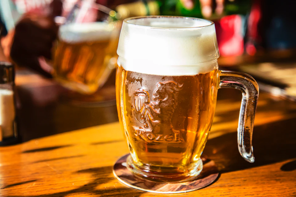 A glass mug of beer with a foamy head, placed on a wooden table, set against a blurred background.