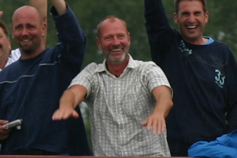 Group of people waving and smiling on the upper deck of a yellow boat with trees in the background.