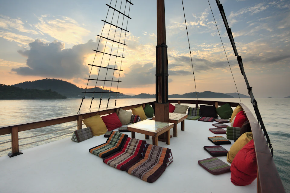 Colorful cushions and low tables on a boat deck with a sunset over calm water and distant hills.