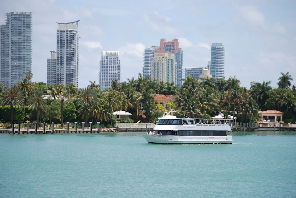 A boat sails on calm water with palm trees and a city skyline featuring tall buildings in the background.
