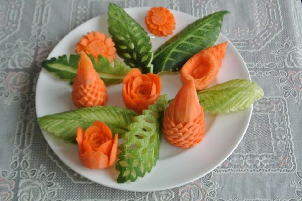 Carved carrots and cucumbers arranged as flowers and leaves on a white plate, placed on a lace tablecloth.