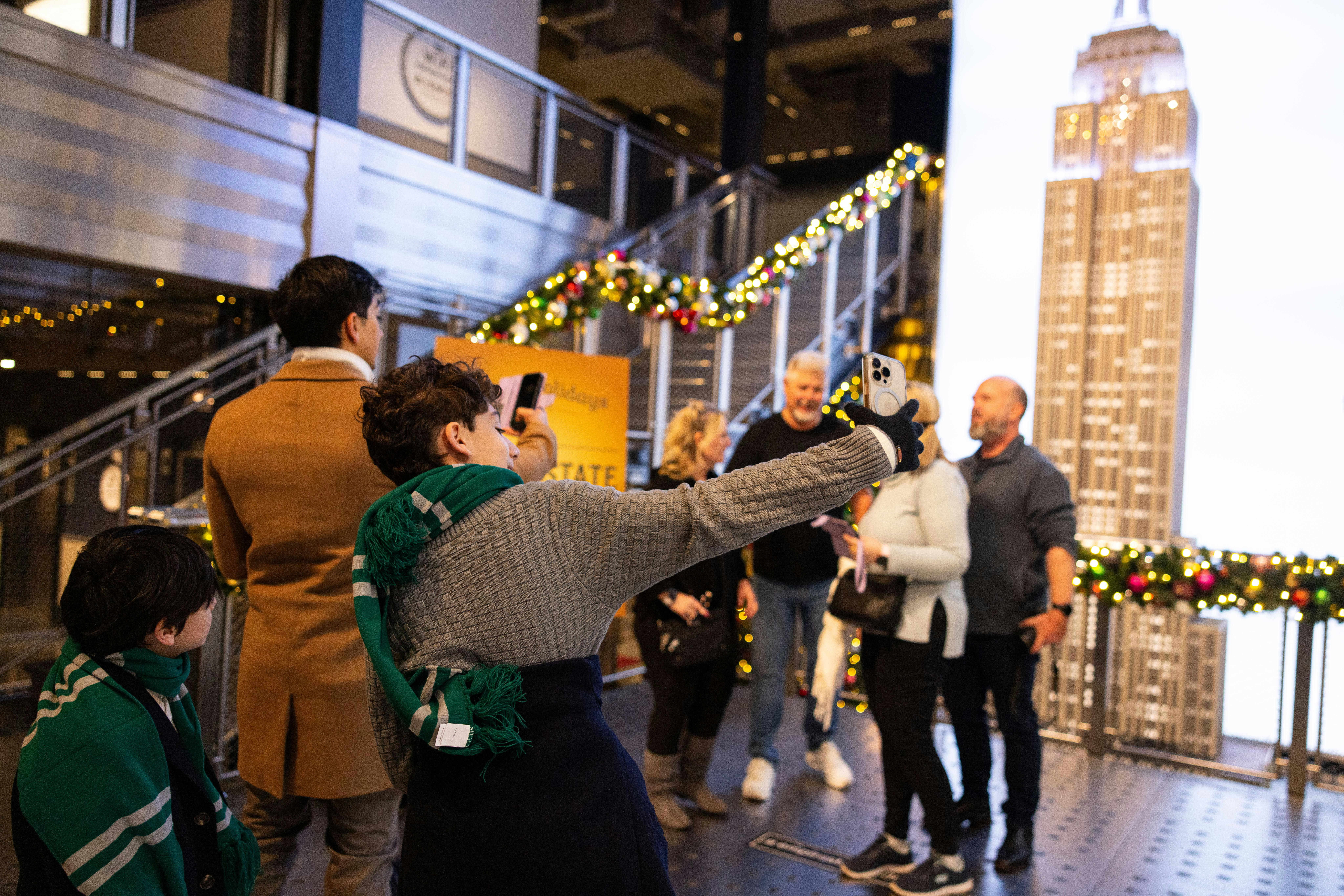 A person taking a selfie near a group of people in front of a lit-up model of the Empire State Building with holiday decorations.
