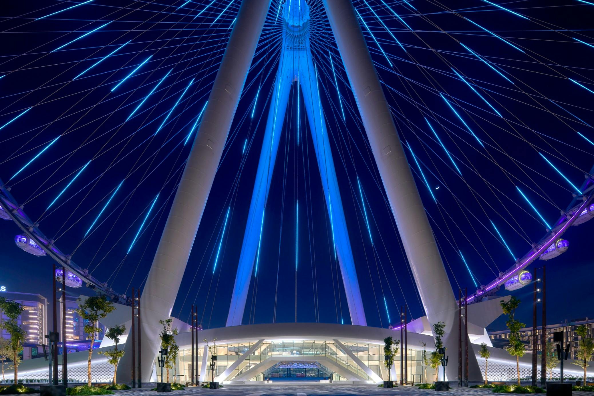 摩天轮在夜空中闪烁着蓝色的灯光，底部有一栋现代化的建筑和树木。