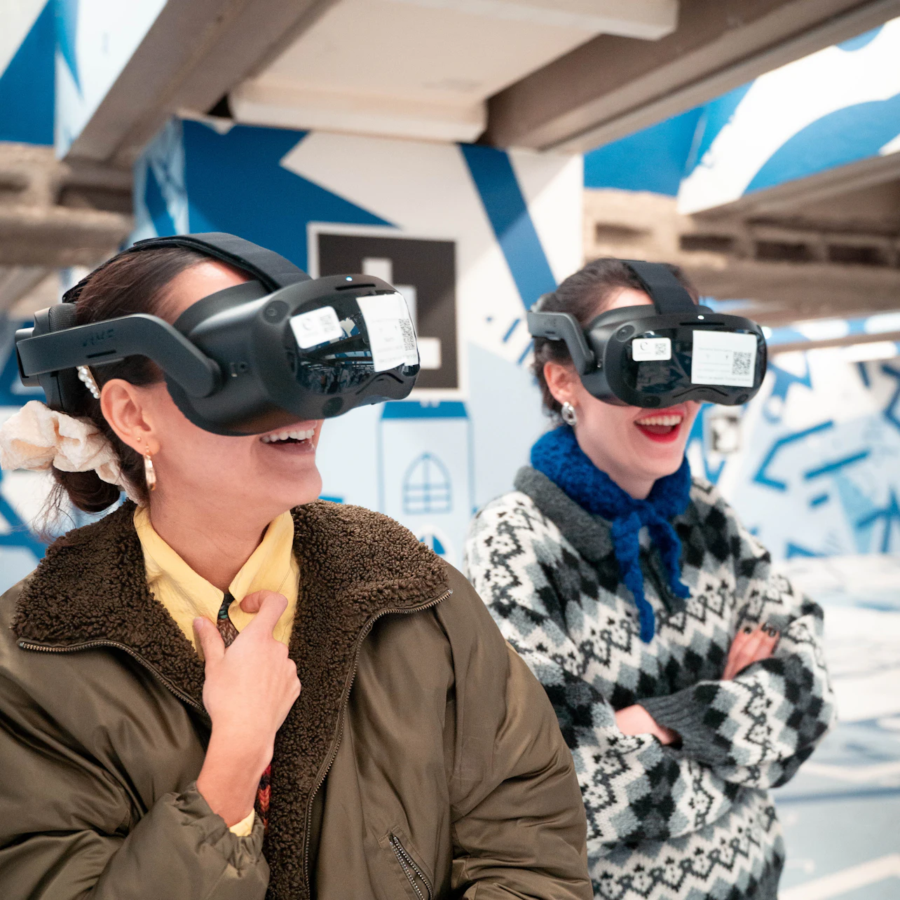 Two women wearing virtual reality headsets, smiling in a futuristic, blue and white setting with abstract patterns.