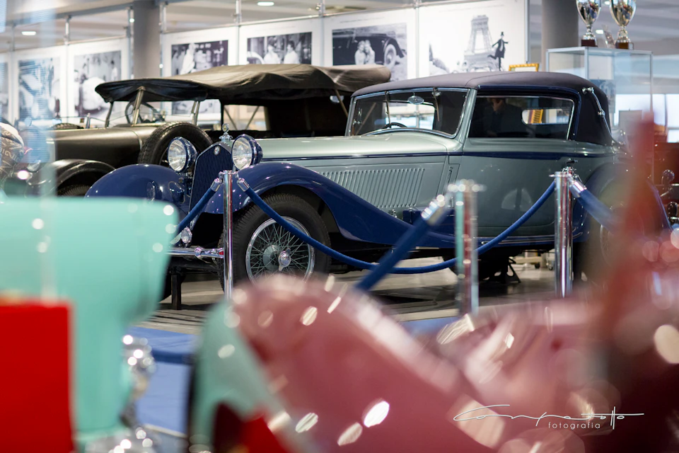 Vintage cars displayed in a showroom, with blue ropes sectioning them off. Historical photos are in the background.